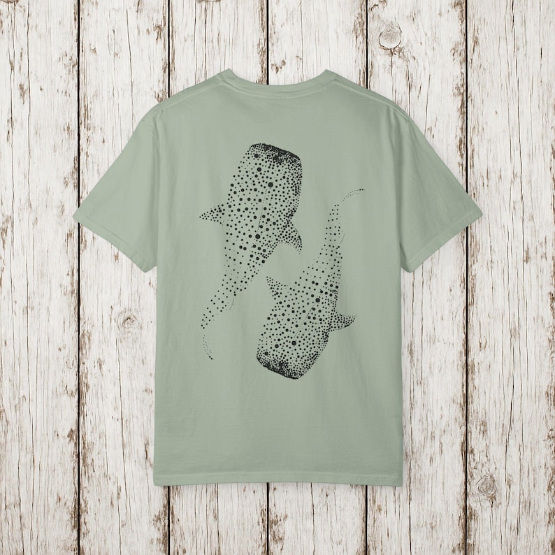 May include: Sage green t-shirt featuring a black illustration of two whale sharks. The whale sharks are depicted with dotted patterns. The t-shirt has short sleeves and a crew neck.