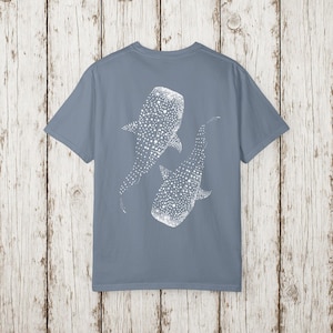May include: A dusty blue t-shirt featuring a white graphic of two whale sharks. The whale sharks are depicted with a pattern of white dots. The t-shirt is made of a soft material and has short sleeves.