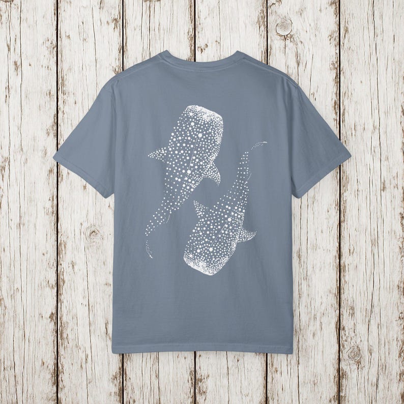 May include: A dusty blue t-shirt featuring a white graphic of two whale sharks. The whale sharks are depicted with a pattern of white dots. The t-shirt is made of a soft material and has short sleeves.