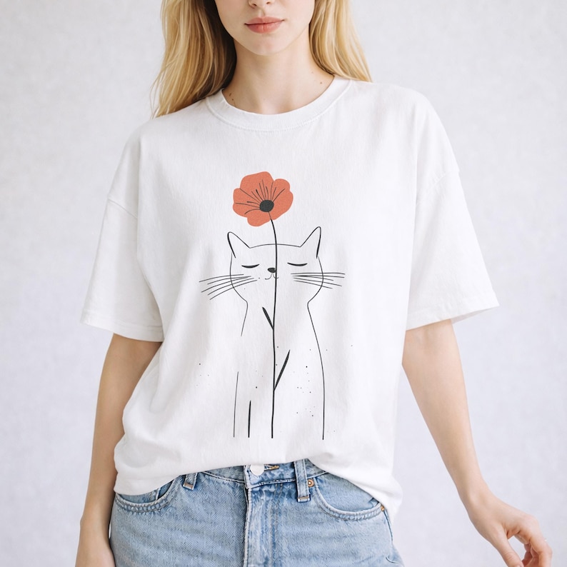 May include: White t-shirt featuring a minimalist cat illustration with a red flower. The cat is drawn with simple black lines, and the flower has a dark center. The shirt is a casual, oversized fit.