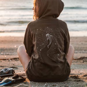 Peut inclure: Un sweat &agrave; capuche marron fonc&eacute; avec un motif de requin-baleine sur le dos, sur fond de ciel &eacute;toil&eacute;. Le sweat &agrave; capuche est port&eacute; par une personne assise sur une plage de sable pr&egrave;s de l'oc&eacute;an.