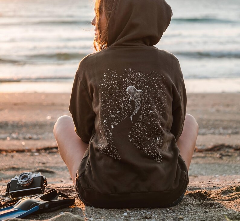 Peut inclure: Un sweat &agrave; capuche marron fonc&eacute; avec un motif de requin-baleine sur le dos, sur fond de ciel &eacute;toil&eacute;. Le sweat &agrave; capuche est port&eacute; par une personne assise sur une plage de sable pr&egrave;s de l'oc&eacute;an.
