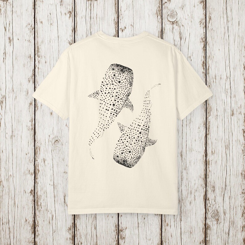 May include: Cream-colored t-shirt featuring a black dotted illustration of two whale sharks. The sharks are depicted swimming in opposite directions. The shirt has short sleeves and a crew neck.
