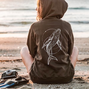 K&ouml;nnte beinhalten: Ein brauner Kapuzenpullover mit einem wei&szlig;en Wal-Design auf dem R&uuml;cken, umgeben von einem gepunkteten Kreis. Eine Vintage-Kamera und blaue Flossen liegen am Strand.