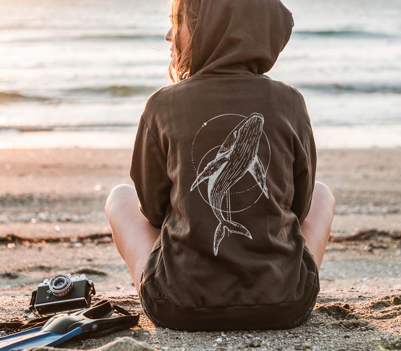 K&ouml;nnte beinhalten: Ein brauner Kapuzenpullover mit einem wei&szlig;en Wal-Design auf dem R&uuml;cken, umgeben von einem gepunkteten Kreis. Eine Vintage-Kamera und blaue Flossen liegen am Strand.