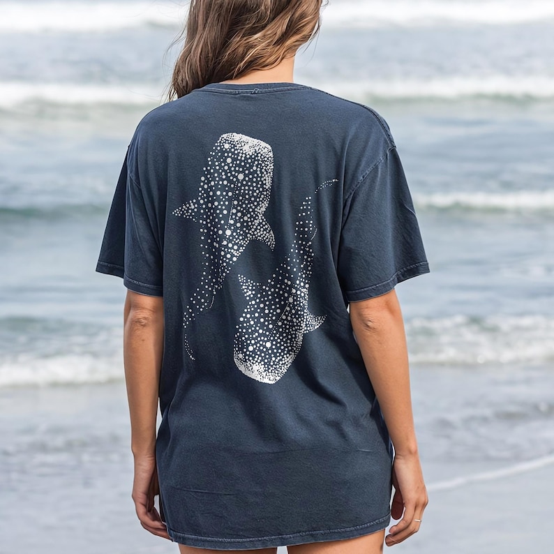May include: Dark navy blue t-shirt featuring a white graphic of two whale sharks. The design is on the back of the shirt. The shirt is made of a soft material and has short sleeves. The background is a blurred view of the ocean.