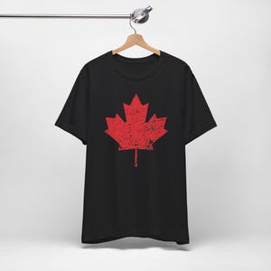 Pode incluir: Uma t-shirt preta de manga curta com um grande gr&aacute;fico central de uma folha de &aacute;cer vermelha. O design da folha de &aacute;cer apresenta uma textura desgastada e envelhecida em toda a sua superf&iacute;cie. A pe&ccedil;a &eacute; exibida num cabide de madeira.