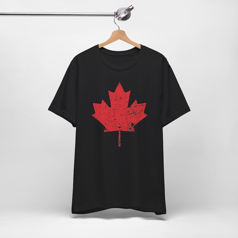 Pode incluir: Uma t-shirt preta de manga curta com um grande gr&aacute;fico central de uma folha de &aacute;cer vermelha. O design da folha de &aacute;cer apresenta uma textura desgastada e envelhecida em toda a sua superf&iacute;cie. A pe&ccedil;a &eacute; exibida num cabide de madeira.