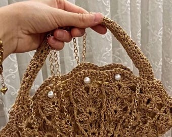 Könnte beinhalten: Eine braune gehäkelte Handtasche mit einer goldenen Kettenkette und Perlenakzenten.