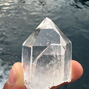 Op de afbeelding: Een heldere kwartskristalpunt met een dubbele beëindiging. De kristal is gefacetteerd en heeft een natuurlijke, ruwe rand. De kristal wordt in een hand gehouden.