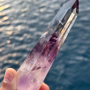 May include: A large, clear crystal point with a purple amethyst tip. The crystal is pointed at the top and has a flat base. The crystal is held in a hand against a blue water background.