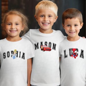 May include: Three children wearing white t-shirts with personalized designs. The shirts feature names in a collegiate font, with accompanying graphics: Cinderella, Lightning McQueen, and Spider-Man. The children are smiling and posing for the camera.