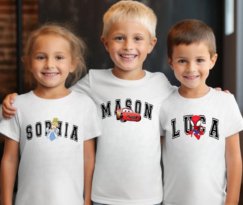 May include: Three children wearing white t-shirts with personalized designs. The shirts feature names in a collegiate font, with accompanying graphics: Cinderella, Lightning McQueen, and Spider-Man. The children are smiling and posing for the camera.