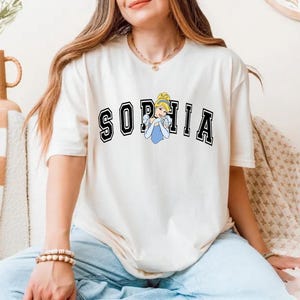 May include: Cream-colored t-shirt with the name "SOPHIA" in black block letters with a white outline. A cartoon image of Cinderella in a blue dress is in the center. The shirt is worn with blue jeans.