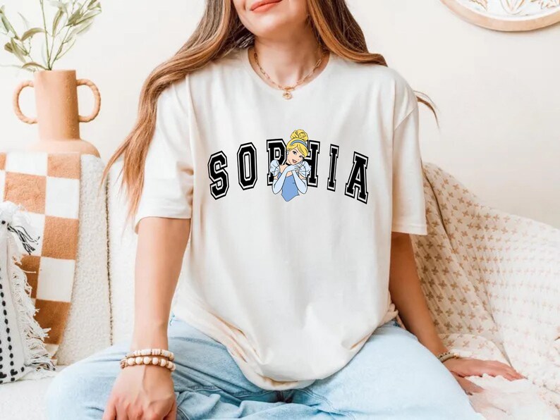 May include: Cream-colored t-shirt with the name "SOPHIA" in black block letters with a white outline. A cartoon image of Cinderella in a blue dress is in the center. The shirt is worn with blue jeans.