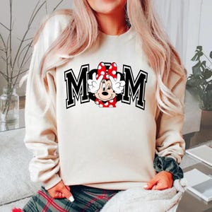 May include: Cream-colored sweatshirt with the word "MOM" in black block letters with white trim. A Minnie Mouse graphic is in the center, wearing a red polka dot bow and making a peace sign with her hands. The sweatshirt is long-sleeved.