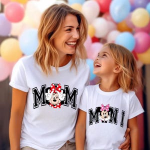 May include: Two white t-shirts with matching designs. One shirt says "MOM" with a Minnie Mouse graphic, and the other says "MINI" with a Minnie Mouse graphic. The shirts are worn by a woman and a child, smiling at each other.