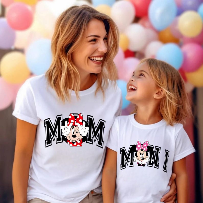 May include: Two white t-shirts with matching designs. One shirt says "MOM" with a Minnie Mouse graphic, and the other says "MINI" with a Minnie Mouse graphic. The shirts are worn by a woman and a child, smiling at each other.