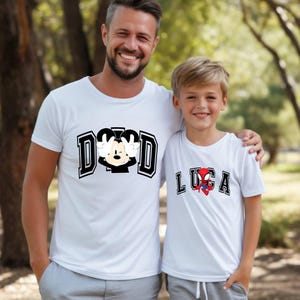 May include: Two white t-shirts with graphic designs. One shirt says "DAD" with a Mickey Mouse design. The other shirt says "LUCA" with a Spider-Man design. Both shirts are worn by a father and son.