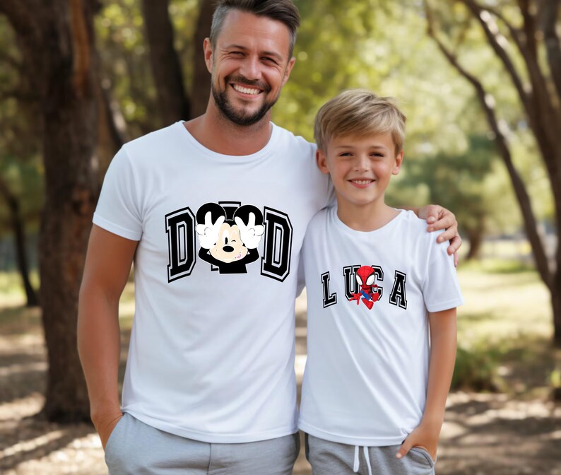 May include: Two white t-shirts with graphic designs. One shirt says "DAD" with a Mickey Mouse design. The other shirt says "LUCA" with a Spider-Man design. Both shirts are worn by a father and son.