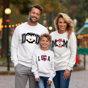 May include: Three people wearing matching white sweatshirts with graphic designs. The sweatshirts feature the words "DAD", "MOM", and "LUCA" with cartoon character designs. The family is smiling in an outdoor setting.