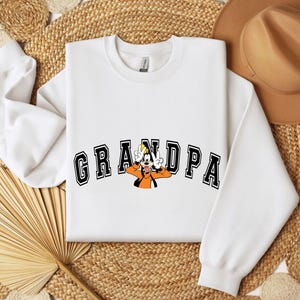 May include: White sweatshirt with the word "GRANDPA" in black block letters with a cartoon character in the center. The cartoon character is wearing an orange shirt and a yellow hat.
