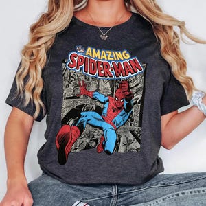 Puede incluir: Camiseta gris oscuro con un gr&aacute;fico de c&oacute;mic de Spider-Man en acci&oacute;n. El gr&aacute;fico incluye el texto "the AMAZING SPIDER-MAN" en amarillo y rojo. La camiseta tiene cuello redondo y mangas cortas.