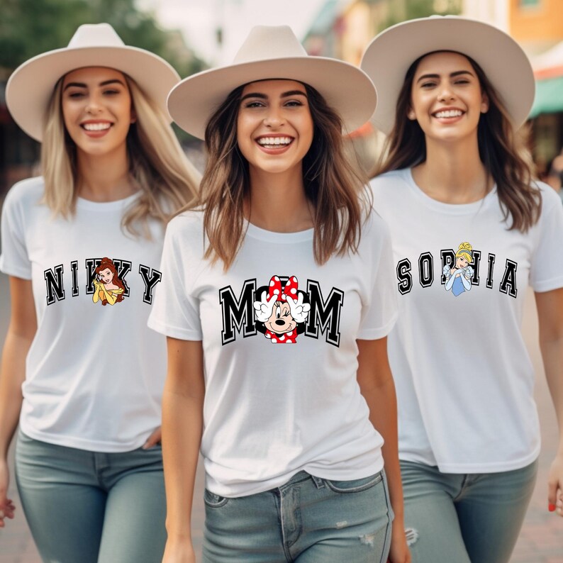 May include: Three women wearing white t-shirts with various designs. One shirt says "NIKKY" with a cartoon image, another says "MOM" with a Minnie Mouse graphic, and the third says "SOPHIA" with a cartoon image. All are wearing hats.