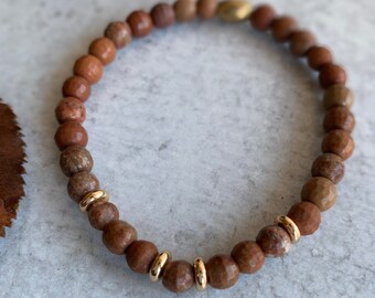 Op de afbeelding: Een elastische armband bestaande uit ronde, gefacetteerde bruine stenen kralen. Het ontwerp bevat verschillende gladde, gepolijste goudkleurige metalen tussenkralen die verspreid zijn over de cirkelvormige opstelling van de aardse, getextureerde bruine edelsteenkralen.