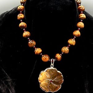 May include: A brown tiger's eye gemstone necklace with a flower-shaped pendant. The pendant is carved from tiger's eye and has a red gemstone in the center.