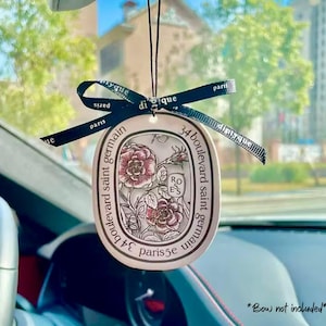 May include: An oval-shaped, off-white car air freshener with a floral design and the text "34 boulevard saint germain paris 5e". A black ribbon is tied at the top. The image is taken inside a car.