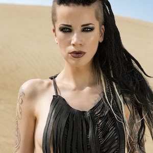 May include: A woman with a shaved head and long black braids wears a black leather fringe top with a cut-out design. She has dark eye makeup and a serious expression.