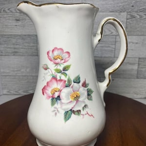 Op de afbeelding: Witte keramische kan met een gouden rand en handvat. De kan heeft een bloemenmotief met roze en witte bloemen en groene bladeren.