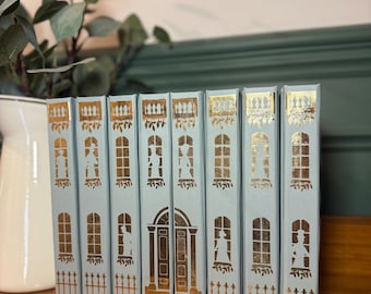 May include: A set of eight blue books with gold accents. Each book features a silhouette of a house with windows and a door. The books are arranged in a row on a wooden shelf.