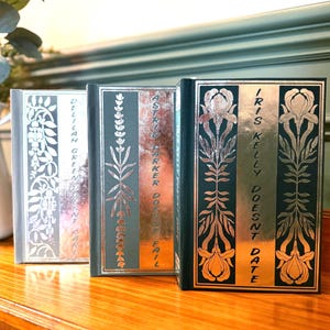 May include: Three hardcover books with decorative silver and gold floral designs. The spines feature titles like "Delilah Green," "Astrid Parker Doesn't Fail," and "Iris Kelly Doesn't Date." The books are displayed on a wooden surface.