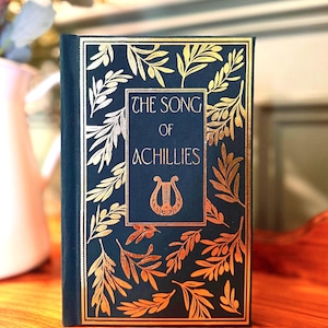 May include: A dark blue hardback book with gold lettering and a gold lyre design on the cover. The title of the book is "The Song of Achilles".