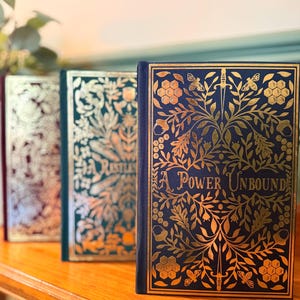 May include: Three hardcover books with gold-embossed floral designs and titles. The book on the right is titled "A Power Unbound".