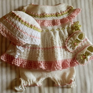 May include: A handmade crochet baby outfit in white, pink, and green. The set includes a dress, shorts, hat, and shoes. The dress and hat have a scalloped edge. The shoes have a flower design.