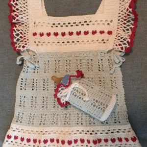 Pode incluir: Um vestido de bebé de crochê em branco e verde claro. O vestido tem corações vermelhos em volta do decote e da bainha. Inclui uma capa de garrafa de crochê.