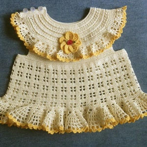 May include: A crocheted baby dress with a yellow and white color scheme. The dress has a ruffled skirt and a matching crocheted collar with a yellow flower.