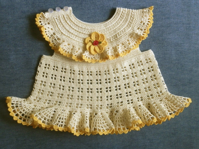 May include: A crocheted baby dress with a yellow and white color scheme. The dress has a ruffled skirt and a matching crocheted collar with a yellow flower.