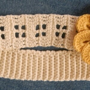 May include: A crocheted headband with a yellow flower accent. The headband is cream colored and the flower has a red center.