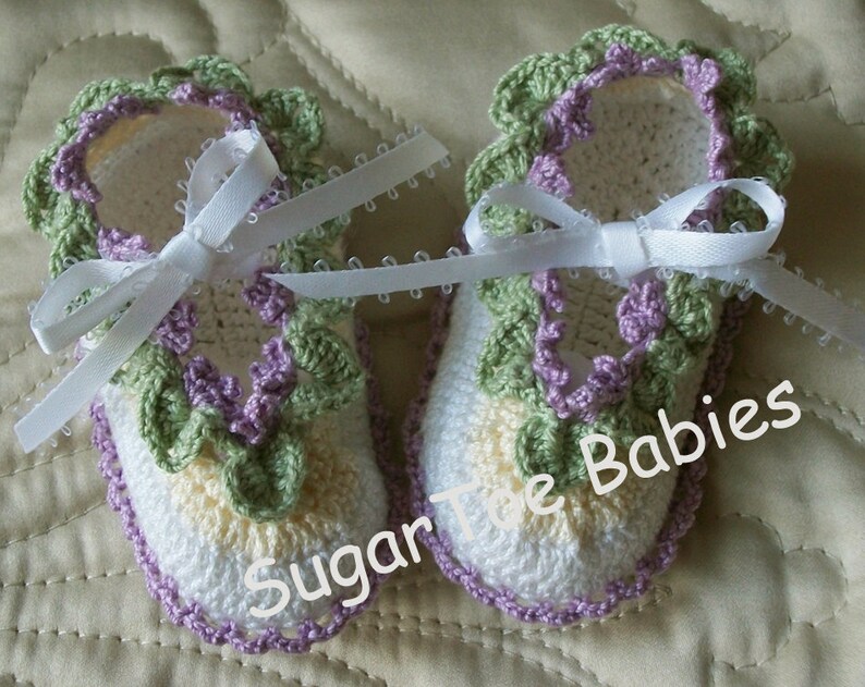 Puede incluir: Un par de botitas de beb&eacute; de ganchillo con un dise&ntilde;o floral blanco, amarillo, verde y morado. Las botitas tienen lazos de cinta de sat&eacute;n blanco. El texto "Sugar Toe Babies" es visible en la imagen.