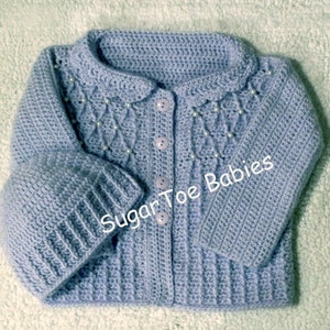 May include: A light blue crocheted baby cardigan with pearl embellishments and matching hat. The cardigan has a button closure and a scalloped collar. The hat is crocheted in the same stitch as the cardigan. The text "Sugar Toe Babies" is visible on the cardigan.