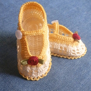 May include: A pair of crocheted baby shoes in yellow and white with a red rose embellishment. The shoes have a simple design and are perfect for a baby girl.