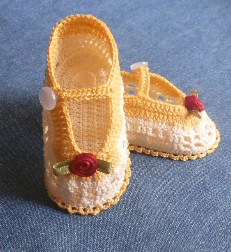 May include: A pair of crocheted baby shoes in yellow and white with a red rose embellishment. The shoes have a simple design and are perfect for a baby girl.