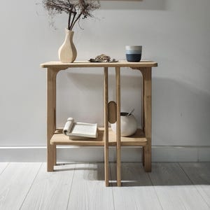 May include: A light wood side table with a framed artwork depicting dancing figures. A vase with dried branches, a small ceramic bowl, and a mug are on the table. A book is on the lower shelf. The artwork and decor create a minimalist aesthetic.