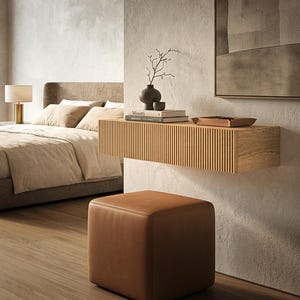 May include: A bedroom featuring a low-profile bed with beige bedding, a floating light wood wall-mounted shelf with vertical slats, and a square tan leather ottoman. The room includes a bedside lamp and a large abstract wall art piece.