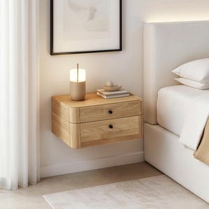 Puede incluir: Una mesita de noche flotante de madera con dos cajones y pomos negros. Una lámpara beige, libros y un cuenco decorativo están encima. La mesita está junto a una cama con ropa de cama blanca y una manta beige. Habitación de diseño moderno.