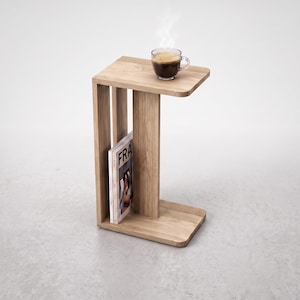 May include: A C-shaped side table crafted from light-toned wood with a smooth grain finish. The table features a flat top surface holding a glass mug of dark coffee with steam rising, and a vertical storage slot containing a magazine titled FRA.